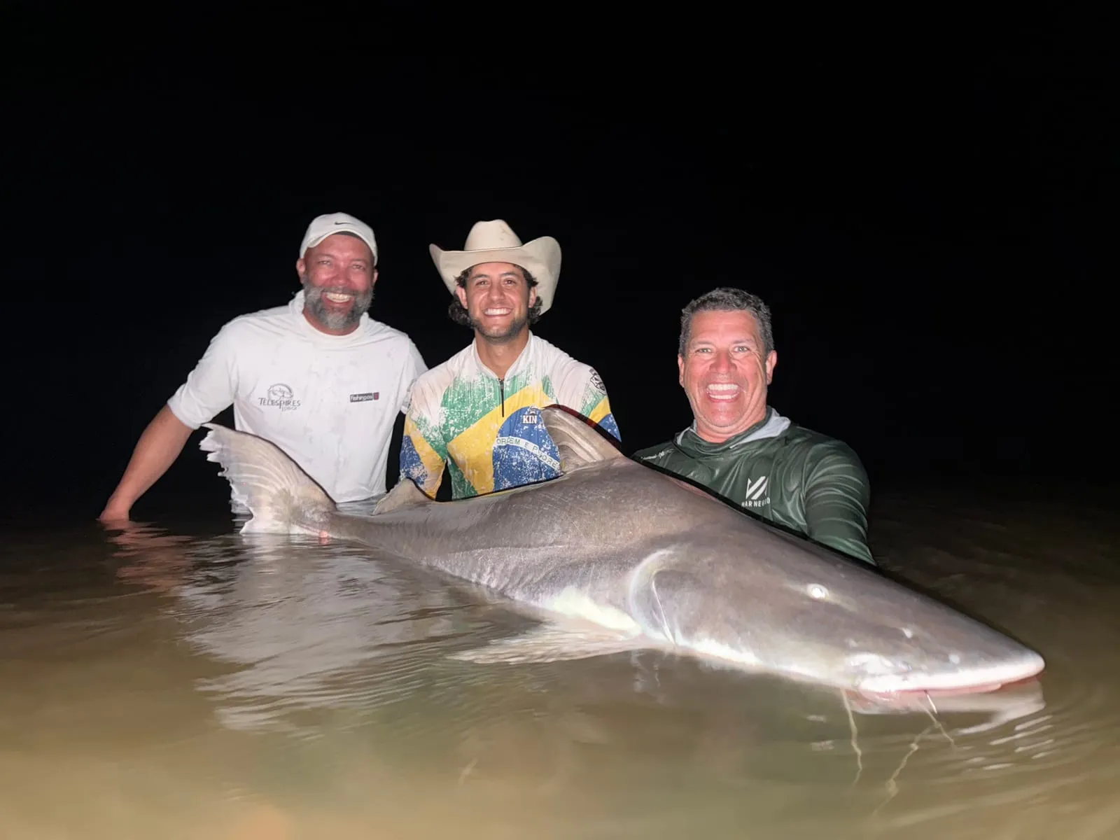 Três pescadores à noite seguram uma piraíba dentro do rio