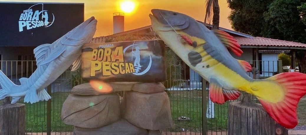 Placa da Pousada Bora Pescar entre duas esculturas de peixes, ao pôr do sol