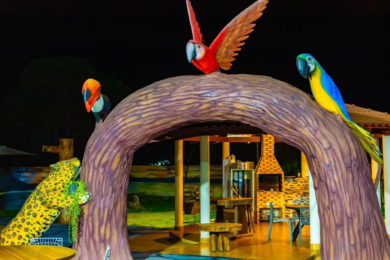 Escultura de tronco com araras e onça, com a área de convivência iluminada ao fundo