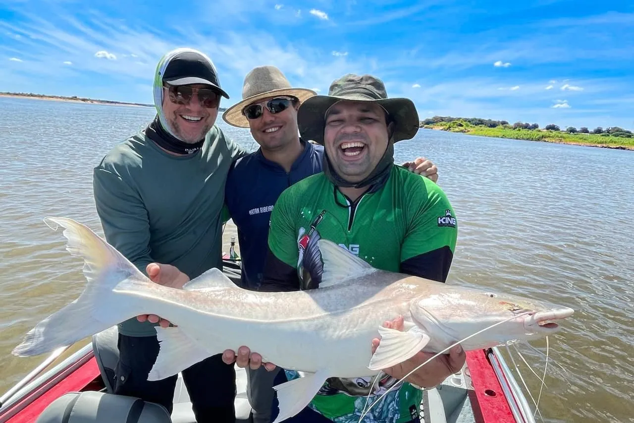 Três pescadores no barco seguram um peixe de couro claro e comprido